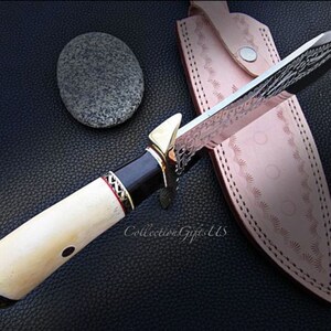 May include: A white-handled knife with a black and gold handle and a silver blade with intricate patterns. The blade is partially inserted into a brown leather sheath with decorative stitching.