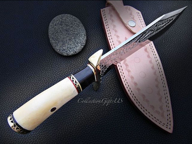 May include: A white-handled knife with a black and gold handle and a silver blade with intricate patterns. The blade is partially inserted into a brown leather sheath with decorative stitching.