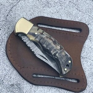 May include: A folding pocket knife with a patterned handle and a partially serrated blade rests on a brown leather sheath. The knife features a gold-colored bolster and rivets. The sheath has a belt loop.