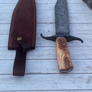 Pu&ograve; includere: Un coltello da caccia artigianale con manico in legno marrone e lama in acciaio damasco. Il coltello viene fornito con un fodero in pelle marrone.