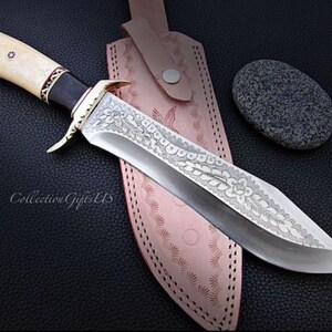 May include: A large, silver knife with a bone handle and intricate floral engravings on the blade. The knife is resting on a brown leather sheath with a decorative stitching pattern. The knife is accompanied by a small, gray stone.