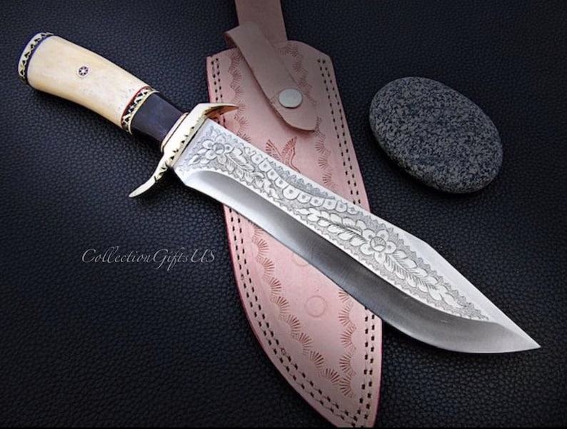 May include: A large, silver knife with a bone handle and intricate floral engravings on the blade. The knife is resting on a brown leather sheath with a decorative stitching pattern. The knife is accompanied by a small, gray stone.