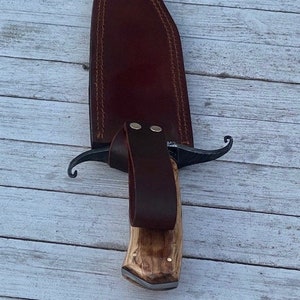 Pu&ograve; includere: Un fodero in pelle marrone con un coltello con manico in legno. Il coltello ha una lama in metallo nero e una guardia in metallo decorativa.