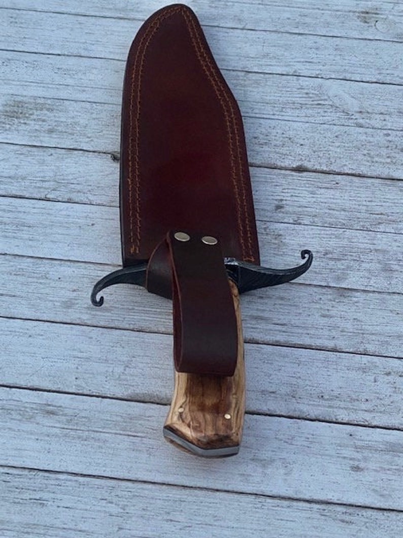 Pu&ograve; includere: Un fodero in pelle marrone con un coltello con manico in legno. Il coltello ha una lama in metallo nero e una guardia in metallo decorativa.