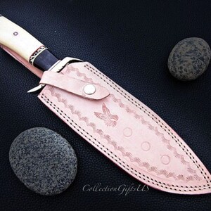 May include: A leather sheath with a decorative design and a white handle knife. The sheath is pink with a white stitching and a bird design. The knife has a black handle with white accents.