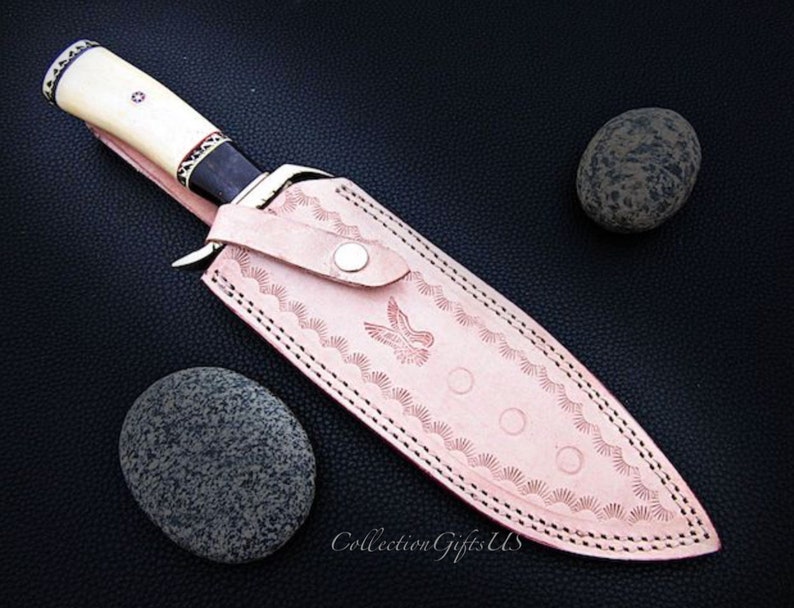 May include: A leather sheath with a decorative design and a white handle knife. The sheath is pink with a white stitching and a bird design. The knife has a black handle with white accents.