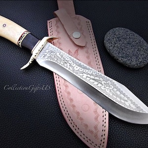 May include: A large, silver knife with a bone handle and intricate floral engravings on the blade. The knife is resting on a brown leather sheath with decorative stitching.