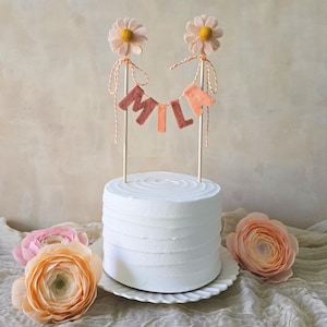 May include: A white frosted cake on a white plate, decorated with a banner that spells out "MILF" and two daisy flower toppers. The cake is surrounded by peach-colored flowers on a neutral background.