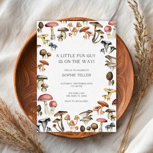 May include: A baby shower invitation featuring a watercolor design of various mushrooms. The invitation is surrounded by a border of mushroom illustrations and includes the text "A LITTLE FUN GUY IS ON THE WAY!" and event details.