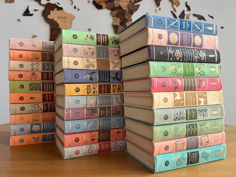 May include: A collection of vintage hardback books with colourful covers and gold lettering. The books are stacked in two piles on a wooden surface. The books have titles such as "The Life of a Bee" and "The Story of a Tree".