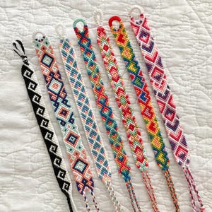 May include: A set of eight colorful friendship bracelets with geometric patterns. The bracelets are made with colorful embroidery floss and have a variety of different designs.