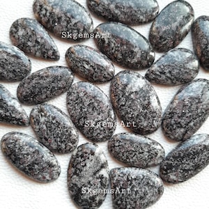 Op de afbeelding: Een verzameling van 15 ovale, gepolijste, zwart-witte edelsteen cabochons met rode vlekken. De stenen zijn willekeurig gerangschikt op een wit oppervlak. SkgemsArt is op de afbeelding gedrukt.