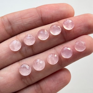 Könnte beinhalten: Zehn facettierte Rosenquarz-Edelsteine in verschiedenen Größen, auf einer Hand präsentiert. Die Edelsteine sind hellrosa und haben eine glatte, polierte Oberfläche.