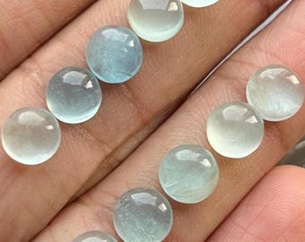 May include: A collection of translucent, round gemstones in varying shades of pale blue and clear. The polished stones are displayed against a neutral background, showcasing their smooth, reflective surfaces. These cabochons are ideal for jewelry making.
