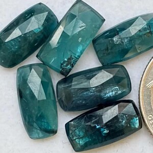 May include: Six rectangular, faceted teal gemstones with a polished finish, arranged in a cluster next to a United States quarter coin for scale. Each stone displays a deep blue-green hue with internal inclusions and geometric cut surfaces.