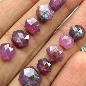 May include: A selection of faceted, deep pink gemstones with a hexagonal form. The gems have a polished, reflective surface, and are displayed against a neutral backdrop. The gemstones vary in size and colour intensity.