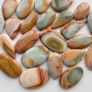 May include: A selection of polished, oval gemstones in shades of tan, brown, and grey. The stones have a smooth, glossy finish and display natural patterns. Some stones have streaks of red and orange. The image includes the text "SkgemsArt".