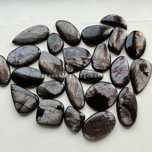 May include: A collection of polished black gemstones with a reflective, iridescent sheen. The stones vary in shape, from oval to teardrop, and are arranged on a light-colored surface. The gemstones have a metallic appearance.