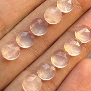 Könnte beinhalten: Eine Sammlung facettierter, runder Rosenquarz-Edelsteine. Die Steine haben eine zarte rosa Farbe und wirken durchscheinend. Die Facetten fangen das Licht ein und erzeugen einen funkelnden Effekt. Die Edelsteine werden vor einem hellen Hintergrund präsentiert.