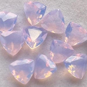 Op de afbeelding: Een verzameling van tien gefacetteerde, driehoekige edelstenen. De edelstenen hebben een zacht, opalescent uiterlijk met een mix van lichtroze en lavendelkleurige tinten. Het licht vangt de facetten op en creëert een glinsterend effect.
