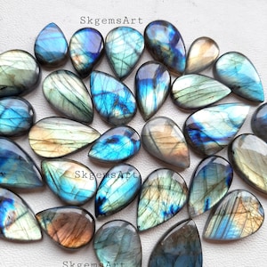 Può includere: Una collezione di 20 cabochon di labradorite a forma di goccia in varie tonalità di blu, verde e oro. Le pietre sono disposte in modo casuale su una superficie bianca. SkgemsArt è scritto tre volte sulla superficie.