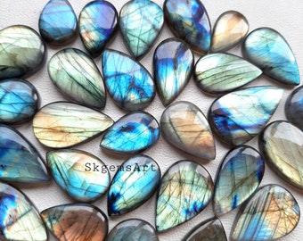 May include: A collection of 20 teardrop-shaped labradorite gemstone cabochons in various shades of blue, green, and gold. The stones are arranged in a random pattern on a white surface. SkgemsArt is written three times on the surface.