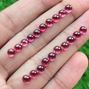 May include: A collection of 18 round, pink gemstones, likely rubies, arranged in two rows on a hand. The gemstones are smooth and polished, with a slight shine.
