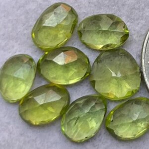 Könnte beinhalten: Eine Ansammlung facettierter, ovalförmiger Peridot-Edelsteine. Die Edelsteine haben eine transparente, hellgrüne Farbe mit mehreren Facetten, die das Licht einfangen. Die Steine liegen auf einer hellen Oberfläche, eine Münze ist teilweise sichtbar.