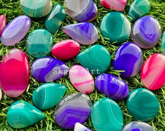 May include: Assortment of agate cabochons in various colors and patterns, including green, pink, purple, and white. The cabochons are arranged on a green grassy surface. The text "SkgemsArt" is visible in the center of the image.