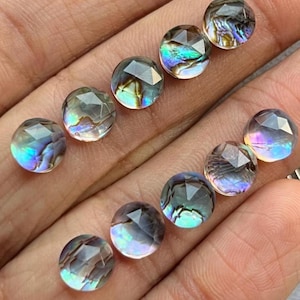 Puede incluir: Una colección de gemas redondas facetadas con patrones iridiscentes, similares a los de la abulón. Las gemas muestran una gama de colores, incluyendo azules, verdes y púrpuras, y se sostienen en una mano. Las gemas son probablemente para hacer joyas.