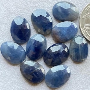 Könnte beinhalten: Eine Ansammlung ovalförmiger, facettierter blauer Saphire. Die Edelsteine variieren in Blautönen, von hell bis tief Indigo. Die Facetten fangen das Licht ein und erzeugen einen funkelnden Effekt. Eine Münze dient als Größenvergleich.