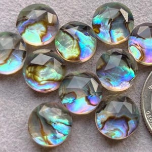 Op de afbeelding: Een verzameling iriserende abalone schelp cabochons met gefacetteerde oppervlakken. De cabochons tonen een reeks kleuren, waaronder blauw, groen en paars. Een munt is inbegrepen ter referentie van de grootte.