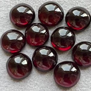 May include: A collection of ten polished, round, dark red gemstones. The gems have a smooth, reflective surface, with a slight gradient of color from dark to light. The gemstones are arranged on a light gray surface.