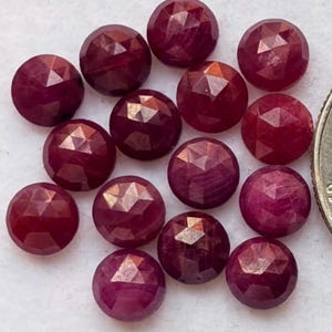 Puede incluir: Un conjunto de trece piedras preciosas redondas, facetadas y de color rojo intenso dispuestas en un grupo. Cada piedra presenta una superficie geométrica de múltiples facetas que refleja la luz, creando un aspecto brillante. Una parte de una moneda metálica es visible a la derecha.