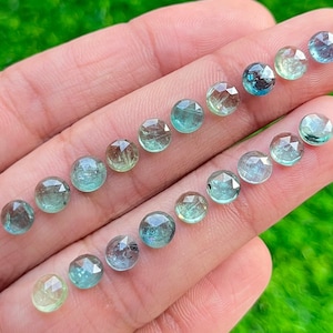 May include: A collection of 18 faceted gemstone beads in shades of green and blue. The beads are arranged in two rows on a hand.