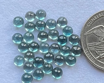 May include: A collection of 25 round, teal blue gemstones, each approximately 6mm in diameter. The gemstones are scattered on a white surface, next to a US quarter for scale.