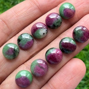 May include: A collection of polished, round gemstones. Each stone features a mix of deep red and green hues, with a glossy finish. The gemstones are held in a hand, showcasing their natural colors and textures. These stones are likely used for jewelry or crafts.