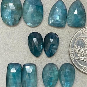May include: An assortment of faceted, teal-colored gemstones in various shapes, including oval, teardrop, and rectangular cuts. A US quarter is included for size comparison. The gemstones have a polished, reflective surface.