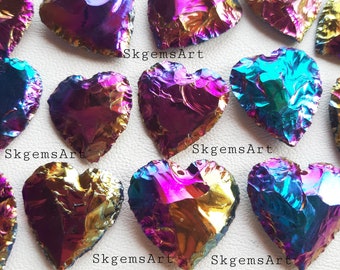 Op de afbeelding: Een verzameling van regenboogkleurige hartvormige stenen met een regenboog van kleuren, waaronder paars, blauw, groen en goud. De stenen zijn in een willekeurig patroon op een wit oppervlak gerangschikt. SkgemsArt staat op de afbeelding.