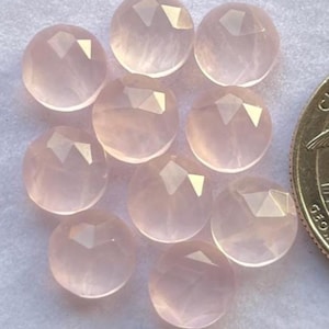 Könnte beinhalten: Neun runde, facettierte Rosenquarz-Edelsteine in einem sanften Rosa. Die Edelsteine haben eine polierte, durchscheinende Oberfläche. Eine Münze dient als Größenvergleich. Durchmesser ca. 2.5 cm.