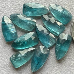 May include: A collection of faceted, teardrop-shaped gemstones in varying shades of teal and aqua. The gems have a polished, reflective surface, with multiple facets catching the light. The gemstones are arranged on a light gray surface.