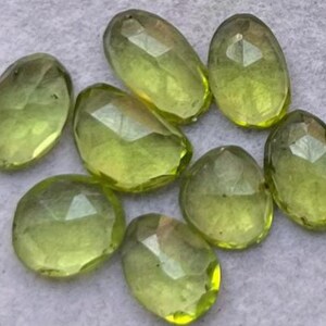Könnte beinhalten: Eine Sammlung facettierter, oval geformter Edelsteine in Olivgrün- und Gelbtönen. Die polierten Steine haben ein durchscheinendes Aussehen, wobei mehrere Facetten das Licht einfangen. Die Steine sind wahrscheinlich für die Schmuckherstellung bestimmt.
