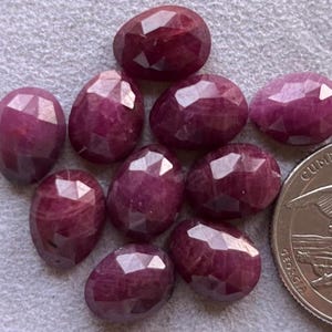 Puede incluir: Una colección de gemas facetadas, de forma ovalada y de color rojo intenso. Las gemas están dispuestas sobre una superficie gris claro. Se incluye una moneda de veinticinco centavos estadounidense para comparar el tamaño. Las gemas tienen una superficie pulida y reflectante.