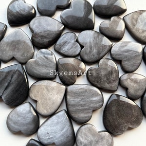 May include: A collection of gray heart-shaped stones with a silvery sheen. The stones are arranged in a pile on a white surface.