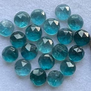 May include: A collection of faceted, round gemstones in varying shades of teal and blue. The gemstones are arranged closely together, showcasing their polished surfaces and geometric cuts. The overall effect is a display of sparkling, translucent stones.