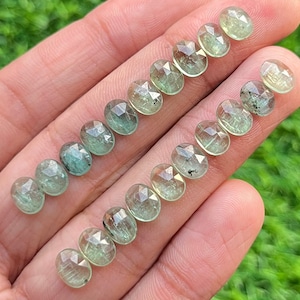 May include: A collection of 18 faceted green gemstones, each with a unique pattern of inclusions. The stones are oval-shaped and have a translucent appearance.