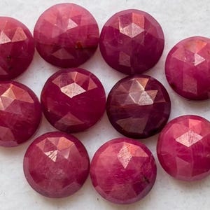 May include: A collection of nine round, faceted, deep pinkish-red gemstones arranged in a cluster. Each stone features a geometric, multi-faceted surface that reflects light, and a portion of a metallic coin is visible on the right edge.