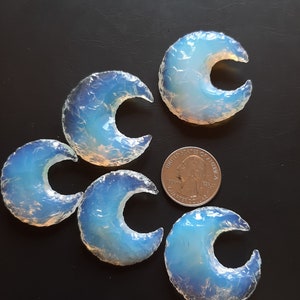 May include: Five iridescent blue and white crescent moon shaped opalite stones on a black background. A US quarter is included for size reference.