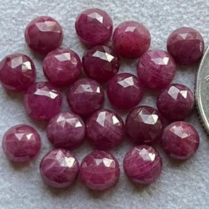 May include: A collection of faceted, round, deep red gemstones. The gems have a polished, reflective surface and are scattered on a light gray surface. The gemstones are likely rubies, and are a popular choice for jewelry making.