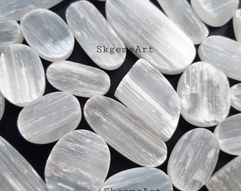 May include: A collection of white selenite crystal stones, each with a smooth, oval shape and a striated texture. The stones are arranged on a black background. The text "SkgemsArt" appears twice in the image.